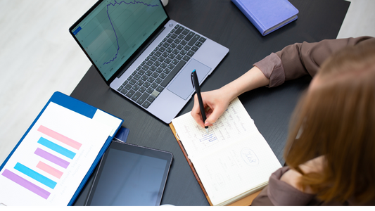 a person writing on a diary or notebook with an open laptop and graph in front of them