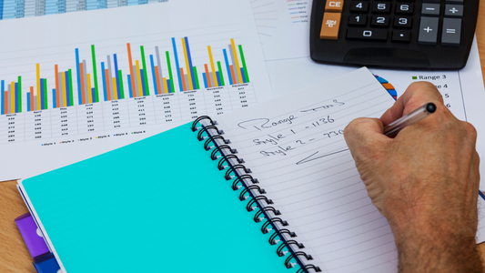 a person writing on a diary with some charts, graphs, and calculator in front of them