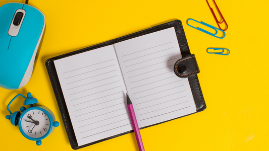 an open diary notebook with a pen, computer mouse, clock, and paper clips surrounding it, on a yellow backdrop