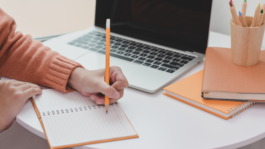 a person writing on a notebook or journal with an opened laptop in front of them